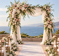Outdoor coastal wedding ceremony arch fully decorated with blush and ivory roses, white flowers and trailing greenery framing an empty aisle