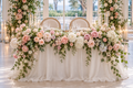 Long sweetheart table for two with blush and ivory garden roses and white hydrangeas arranged across entire front, trailing greenery, candles, flowing ivory draping and two chairs